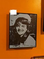 Framed monochrome portrait of Jackie Kennedy smiling, mounted on an orange wall with a black frame.