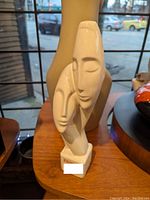 Front and side view of the abstract lovers sculpture on a wooden table showing the stylized intertwined faces and smooth cream finish.