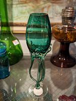 Mid century blown glass vase in teal with unique elongated shape and tubular stem base, presented against a backdrop of other glassware.