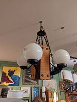 Photo showing a mid century chandelier mounted on ceiling with three white globe glass shades, black metal arms, central wooden block, and black metal chains.