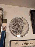 Photo of a circular plaster wall art with a detailed moon face design mounted on a white wall, partially surrounded by framed pictures.