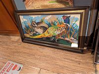 Framed needlepoint bird scene artwork resting against a door and on a wood surface showing the entire piece and frame wear.