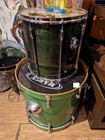 Photo showing pair of drums including dark green floor tom and metallic green bass drum, placed on wooden floor next to decorative painting.