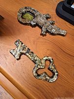 Two resin animal shaped figures on a wooden surface; top one is a crocodile, bottom one is a vintage key design, both with embedded shell or stone mosaic patterns.