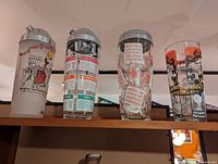 Photo showing four vintage glass bar shakers with colorful printed cocktail and bar-related designs, each with a metal shaker lid and built-in strainer.