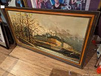 Framed oil painting leaning against the wall showing a detailed urban riverside scene with buildings and a boat, dated mid-20th century and signed.