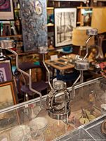 Photo of a silver metal steampunk-style candle holder with five candle platforms arranged on curved arms, displayed on a glass surface in a room with various artworks and furniture.