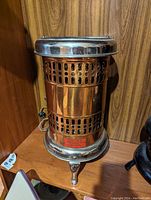 Copper and chrome vintage electric heater with perforated ventilation, standing on three curved legs, displayed on wooden surface.