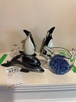 Overview of all six art glass animal figurines on white surface showing two penguins, clear bear, black dolphin, and tropical fish with blue core.