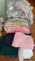 Stack of various blankets and comforters including pink knitted blanket and floral-patterned comforters packed in plastic bags and folded blankets in multiple colors.