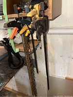 Photo showing large bar clamps and a chain clamp hanging on a wooden rack in a workshop environment.