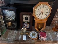 Five clocks grouped together on a brick ledge including three wall clocks, one tabletop clock, and one boxed water clock.