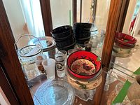 Glass cabinet shelf displaying various dishware items including black mugs, cut glass basket, red and floral serving bowls, frosted glasses, and playing card themed plates.