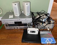 Lot overview showing Panasonic VHS/DVD combo player, JVC stereo cassette deck, Enjoy speakers, wired headphones, stopwatch, and cables on wooden floor.
