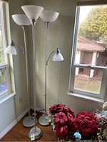 Three silver-tone floor lamps with white shades, two have adjustable reading lamps attached, positioned in a corner near windows with red flowers at base.