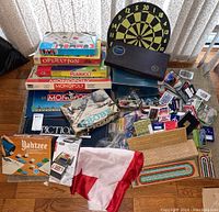 Full lot view depicting multiple board games including Monopoly, Pictionary, Operation, Rummoli, Trivial Pursuit, dartboard, and card decks with game pieces scattered