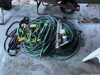 Lot of garden hoses, sprinklers, and bird feeder placed on ground near wheelbarrow and chair showing condition and quantity.