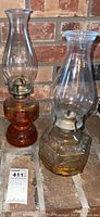 Two vintage hurricane oil lamps with glass chimneys and metal wick holders, showing amber and clear bases with oil inside.