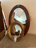 Two mirrors showing their different frames and shapes, positioned with the smaller round gold frame mirror in front of the larger oval wooden framed mirror.