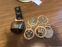 Photo of a brass bell with a black leather strap embossed with a bear and floral detail along with six metal souvenir tags with various designs and text.