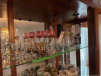 Shelf display of various stemware including clear wine glasses, stemless glasses, and red etched wine glasses arranged in rows on a glass shelf.