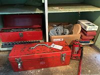 Red metal tool boxes, socket set, hydraulic bottle jack, and a box with assorted hardware and tools on worn shelves in a shed.