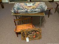 Photo showing all items together: carved colonial wall art plaque, child's wooden bench, folding TV tray table, and rolled decorative rug on top of table.