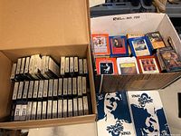Two open boxes filled with 8-track tapes, arranged tightly packed in rows with some tapes in plastic cases and some sealed unopened tapes.