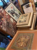 Stack of framed art leaning against each other showing bicycle photograph, abstract tribal painting, ship scene and mosaic panel at front