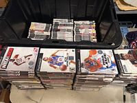 Box containing stacks of PS2 game cases with visible front covers of basketball video games including NBA 2K, NCAA March Madness, and ESPN College Hoops series.
