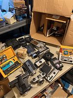 Photo showing a table with various vintage cameras, flashes, Kodak Ektralite 10 box, and camera accessories, some leather cases visible
