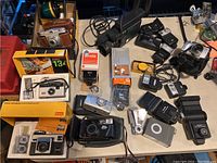 A wide view of the table displaying various vintage cameras including Kodak Instamatic models, flash attachments, straps, and cases.