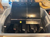 Six PlayStation 3 consoles stacked in a grey bin, top view showing logos and disc slots.