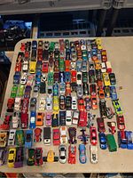 Photo showing approximately 99 vintage Hot Wheels diecast toy cars spread on a table, laid out in organized rows, showing various colors and models.