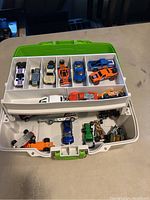 Top view of white and green storage case with vintage Hot Wheels cars arranged in compartments. Cars include varied colors and styles such as white car with racing stripes, gold convertible, orange race car, blue coupe, and others.