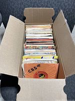 Cardboard box filled with over 130 assorted 45 RPM vinyl records in paper sleeves from various labels including CBS.