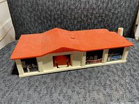 Overall front view of the plastic building playset with orange roof and illustrated windows showing people and cars, central car launch area visible.