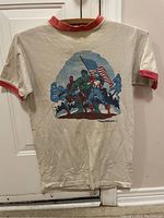 Full front view of off-white short sleeve t-shirt with red collar and sleeve edge ringer detail and a color graphic of Marvel superheroes Spider-Man, Hulk, and Captain America against a smoky blue background.
