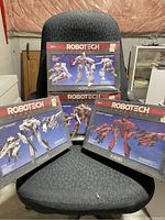 Four Robotech Changers model kit boxes, three standing side by side in front and one resting on top of them, displayed on a dark gray fabric chair. Boxes show vintage design with robot images on front.