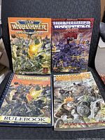 Photo shows four original Warhammer Fantasy books arranged with two standing and two laying flat, depicting detailed fantasy battle scenes on each cover.