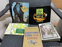 Photo showing five J.R.R. Tolkien books placed on a grey upholstered chair. Books include The Tolkien Scrapbook, The Lord of the Rings boxed set, Journeys of Frodo, Unfinished Tales, and The Silmarillion.