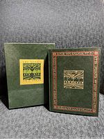 Front view of the dark green hardcover book with gold and red decorative embossed pattern and matching slipcase with illustration.