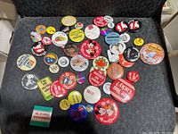 Photo showing a collection of about 40 vintage pins and buttons from the 1970s with varied designs, colors, and text, laid out on a dark fabric surface.