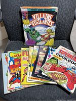 Photo showing 8 comic and role-playing game books spread on a chair, titles include Villains and Vigilantes RPG, Superhero Cookbook, and Mighty Thor Fun Books.