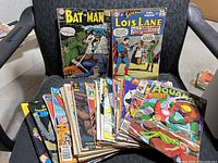 Photo showing a collection of assorted comic books including Batman, Superman, Lois Lane, Aquaman, and various others, laid out on a chair.