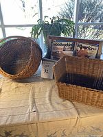 Photo showing a round woven basket, a rectangular woven basket, and a wooden farmhouse tote containing woodworking magazines, all placed on a beige cloth near a window.