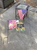 Garden flags displayed on ground with metal stakes; brown wicker table with glass top beside them