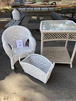 Set of three white wicker patio items outdoors on concrete: armchair, side table with glass top, and basket holder