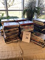 Multiple stacks of over 75 DVDs arranged on a table near a window, showing a variety of DVD cases and titles in original packaging, some opened, some unopened.
