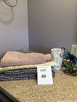 Photo showing three bath rugs stacked in pink, beige, and grey colors on a bathroom counter with the dragonfly night light and two white cup holders nearby.